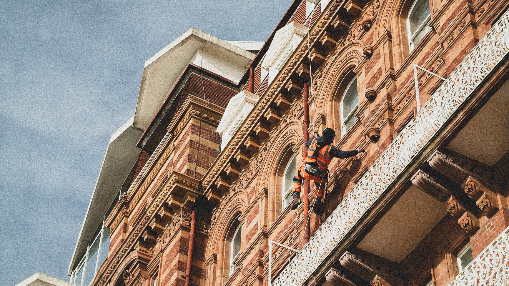 Worker in high-vis gear abseiling on decorative Victorian brick building against a clear blue sky.