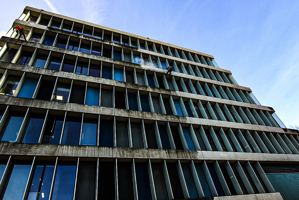 Facade cleaning company in safety gear abseiling down a modern glass building against a clear blue sky.